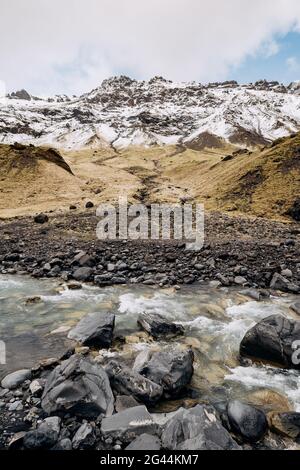 Mountain river at the foot of the mountain with a snow-capped peak. Yellow dry grass on the mountains in May in Iceland. Stock Photo