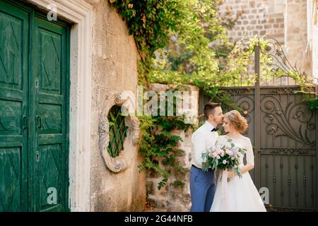 The bride and groom stand hugging near beautiful old green door, the groom kisses the bride on the forehead Stock Photo