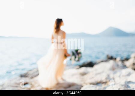 Silhouette of lovely bride in a pastel wedding dress stands on a rock above the sea with a bouquet of flowers in her hands Stock Photo