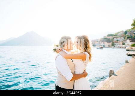 The bride hugs the groom and holds a bouquet in her hands on the pier in the Bay of Kotor Stock Photo