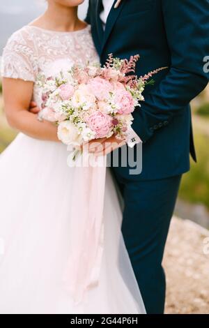 The bride and groom stand hugging and hold the bride's bouquet with delicate pink roses, peonies and astilbe, close-up Stock Photo