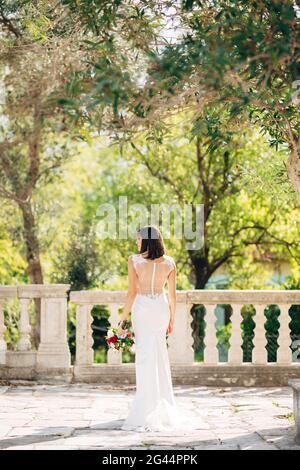 Bride in a white dress with a bouquet of flowers stands with her back on the balcony against the background of green trees Stock Photo