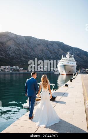 The bride and groom are walking holding hands along the pier near white tourist liner, back view Stock Photo