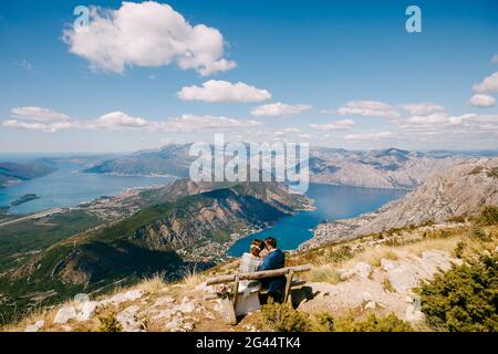 Bride and groom are sitting on a bench and looking at the Kotor bay. Excellent view from the mountains of Montenegro Stock Photo
