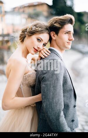 Pensive bride hugs groom from behind while standing on the promenade of Lake Como Stock Photo