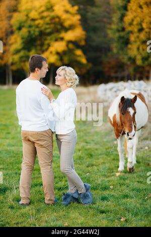 Man hugs a woman while standing on the lawn in the autumn forest. Horse grazing on the lawn Stock Photo