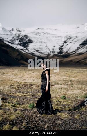 Destination Iceland wedding. A bride in a black dress with a developing train and hair, stands in a field with yellow grass, on Stock Photo