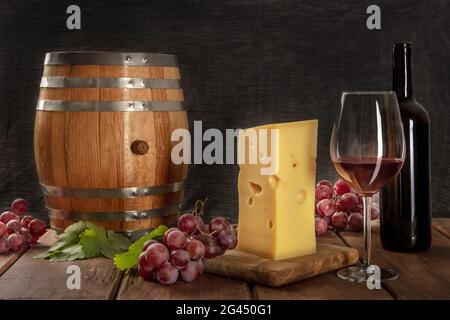 A glass of red wine with a bottle, a wine barrel, a large piece of cheesse, grapes, and vine leaves Stock Photo