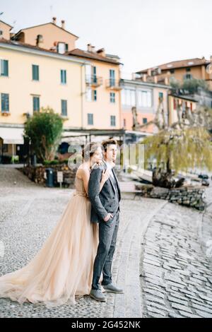 Bride hugs from behind groom while standing on the embankment of Lake Como against the background of old houses Stock Photo