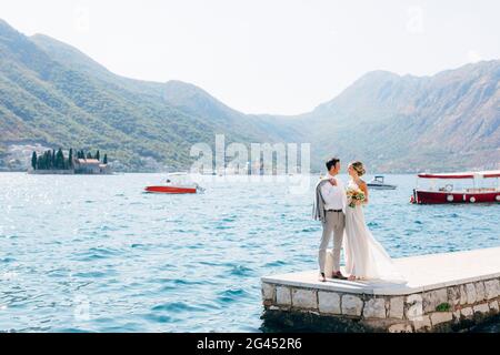 The bride and groom standing side by side on the pier in the Bay of Kotor, islands of Perast are behind them Stock Photo