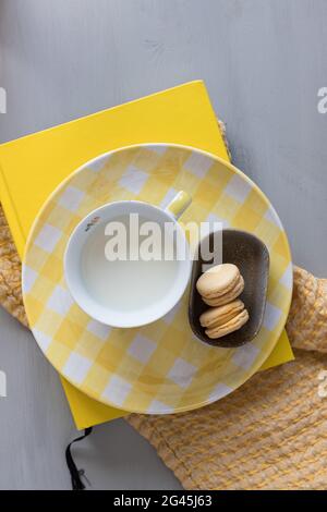 A relaxing single yellow breakfast with two macaroons, a cup of milk and a polka dot dish over an orange cloth on a gray table. Stock Photo