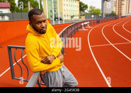 Young pensive handsome african american sportsman runner wearing yellow sportswear looking at camera, having rest after run training ar stadium Stock Photo