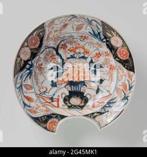 Shaving Bowl With a Flower Vase, Quails and Flower Sprays. Shaving basin of porcelain, painted in underglaze blue and on the glaze red and gold. On the flat a flower vase with peonies and a thriving branch at a fence. A lotus drink on the vase. The edge is divided into six cartouches with alternating two quail in a flowering plant and chrysanthemes on a ground of underglaze blue. The outer wall with two prunus branches. A preen on the bottom. Imari. Stock Photo
