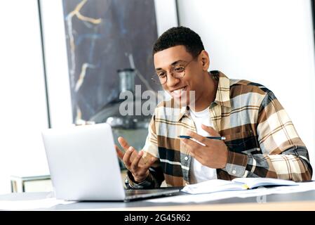 Smart modern latino guy, freelancer, manager or student, sitting at his work desk, using a laptop for online communication, talks over video communication, works or studies remotely, smiling Stock Photo