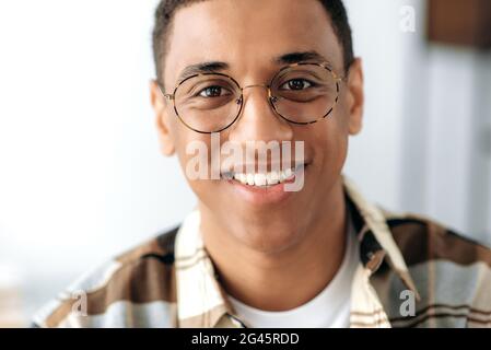 Close-up headshot of a handsome positive millennial hispanic guy with glasses, freelancer or student, standing indoors, looking directly at the camera, smiling friendly Stock Photo