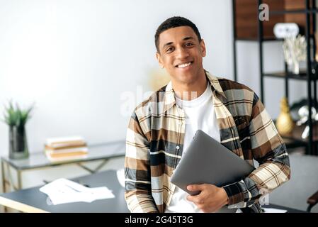 Portrait of a handsome charismatic modern hispanic guy, dressed in stylish shirt, office worker, freelancer or student, holding a laptop, standing near a table, looking at the camera, smiling friendly Stock Photo