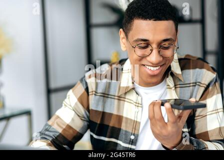 Positive friendly friendly hispanic guy in casual shirt and with glasses, sits at a desk and recording an audio message or talking on smartphone on speakerphone with friend or family, smiling happily Stock Photo