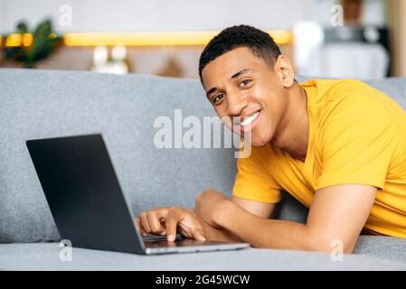 Latino positive guy in a yellow t-shirt, chilling at home, lies on a couch, uses a laptop, chatting online with friends on social networks, looks at the news,looks at camera ,smiles friendly Stock Photo