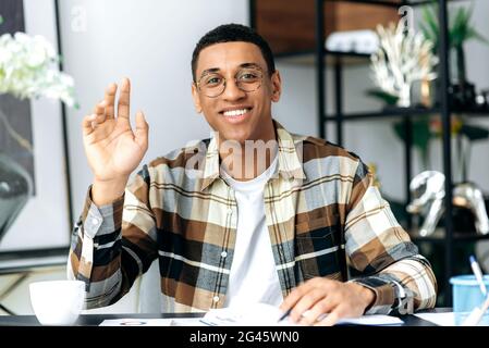 Distant communication. Charismatic friendly hispanic guy in glasses and in a casual shirt, sits at his work desk, looks at the camera, communicates on a video call, greets the interlocutor, smiles Stock Photo