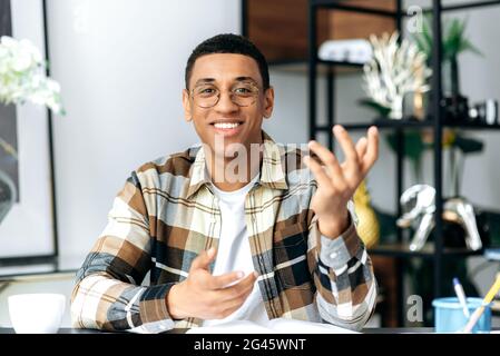 Video conference. Handsome friendly latino guy in glasses and in a casual shirt, sits at his work desk, looks at the camera, communicates on a video call, gesturing with hands, smiles pleasant Stock Photo