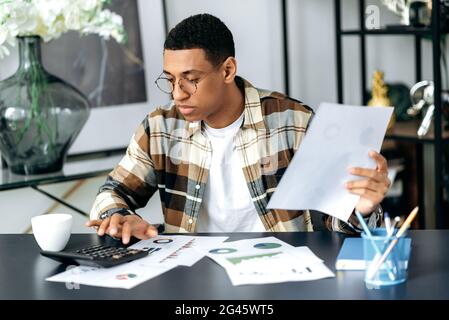 Focused modern hispanic young man with glasses, office worker or freelancer, sitting at his work desk, views graphs, analyzes reports and profit, works with documents in the office Stock Photo