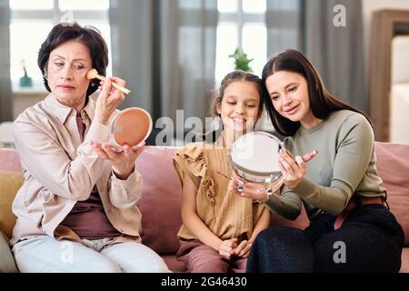 Three females applying makeup while sitting on sofa in living-room Stock Photo