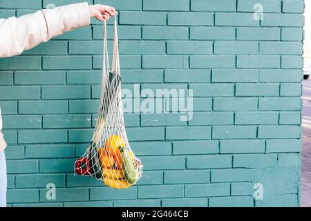Woman hand holding an eco shopping bag with vegetables, fruits in front of green brick background Stock Photo