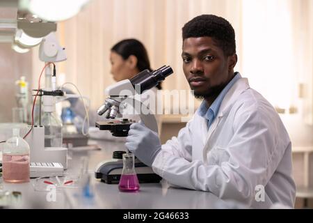 Young African male chemist with microscope sitting by desk in laboratory Stock Photo