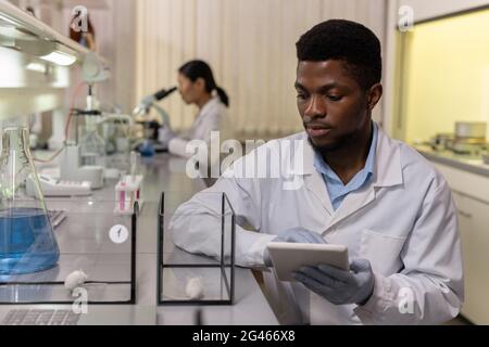 Young scientist of African ethnicity looking at mice in glass boxes and using tablet Stock Photo