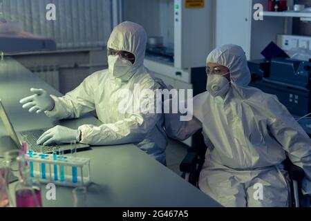 Young scientist in coveralls explaining data on laptop screen to his disabled colleague in wheelchair Stock Photo