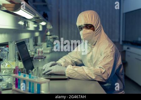 African male scientist in coveralls and protective gloves, eyewear and respirator using laptop Stock Photo