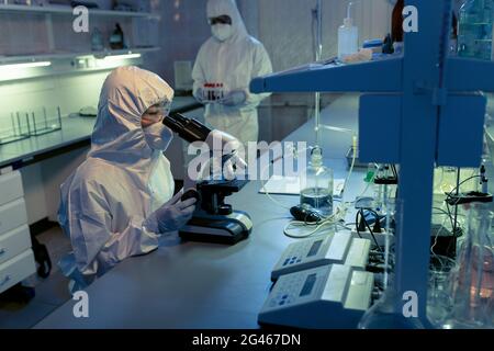 Scientist using microscope while her colleague carrying flasks with medical samples Stock Photo
