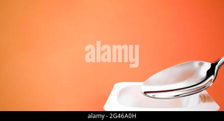 Yogurt cup and silver spoon on orange background, white plastic container with yoghurt cream, fresh dairy product for healthy di Stock Photo