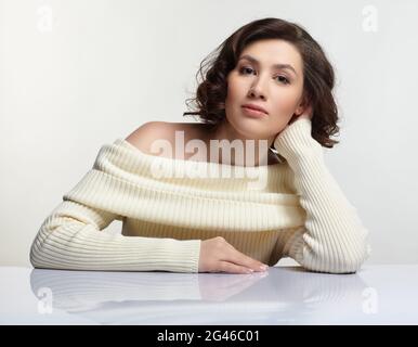 Beautiful brunette woman dressed in large white woolen sweater is sitting at white table. Stock Photo