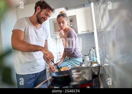 beautiful young caucasian couple having fun in the kitchen. Beardy guy is making breakfast while his female partner tasting. Stock Photo