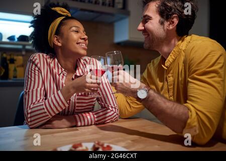 young adult multiethnic couple having toast with glasses of wine, looking each other, smiling. Stock Photo