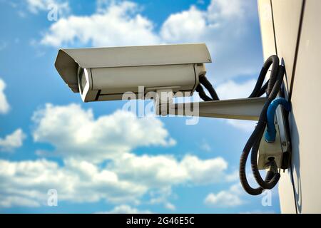 This is a security camera hanging on the wall of a building against a blue sky with clouds on a summer day. Stock Photo