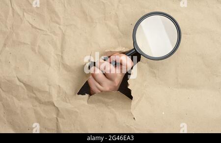 A woman's hand holds a glass magnifier, part of the body sticks out of a hole in brown paper Stock Photo