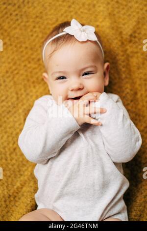 Smiling baby girl with a flower on her head in a white blouse lies on a yellow bedspread with her hands in her mouth Stock Photo