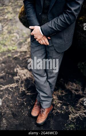 Destination Iceland wedding. Closeup of male legs in gray trousers and leather brown boots. Hands in a jacket hold on to the wri Stock Photo
