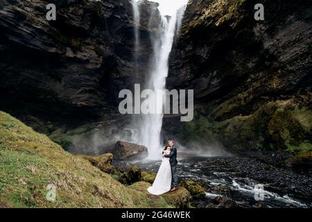 Destination Iceland wedding, near Kvernufoss waterfall. Wedding couple is standing near the waterfall. The groom hugs the bride. Stock Photo