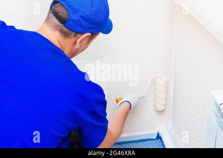 A painter in a cap and gloves paints the wall with a paint roller. Portrait of a young worker in uniform. Stock Photo
