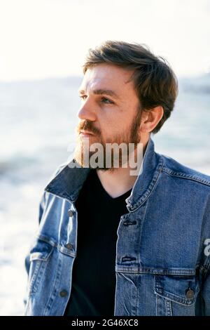 Portrait of a pensive man in a denim jacket looking into the distance against the background of the sea. Close-up Stock Photo