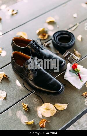 Black shoes of a man with untied laces on a wooden texture with a belt and groom's boutonniere on a white pocket square. Stock Photo