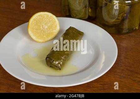 wine leaf roll filled with rice or meat called dolma or sarma in oriental cuisine on a plate with a lemon and more in pickle jars behind Stock Photo