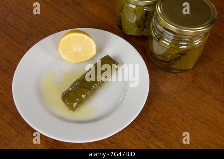 wine leaf roll filled with rice called dolma or sarma in oriental cuisine on a plate with a lemon and more in pickle jars behind Stock Photo