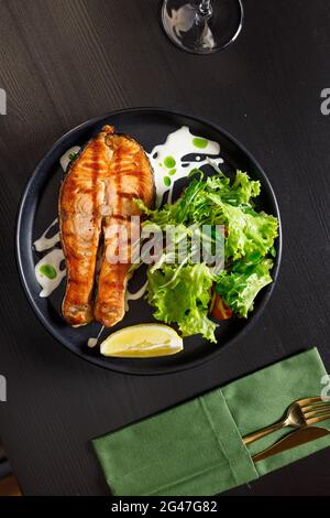 One piece of baked salmon grilled pepper lemon and salt on a black plate with lettuce leaves on a stone background Stock Photo
