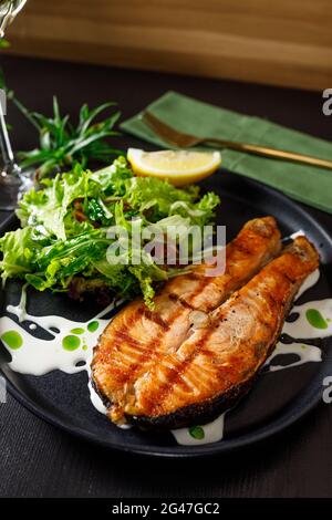 One piece of baked salmon grilled pepper lemon and salt on a black plate with lettuce leaves on a stone background Stock Photo
