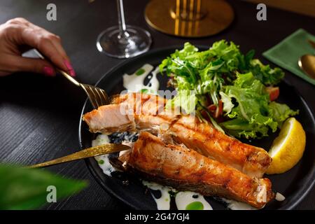 One piece of baked salmon grilled pepper lemon and salt on a black plate with lettuce leaves on a stone background Stock Photo