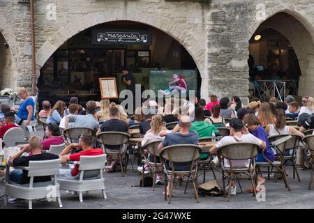 Uzes, Provence, France 06.19.21 Men and women sitting outside a cafe watching a live football match on large screen. Medieval stone arches. drinting. Stock Photo